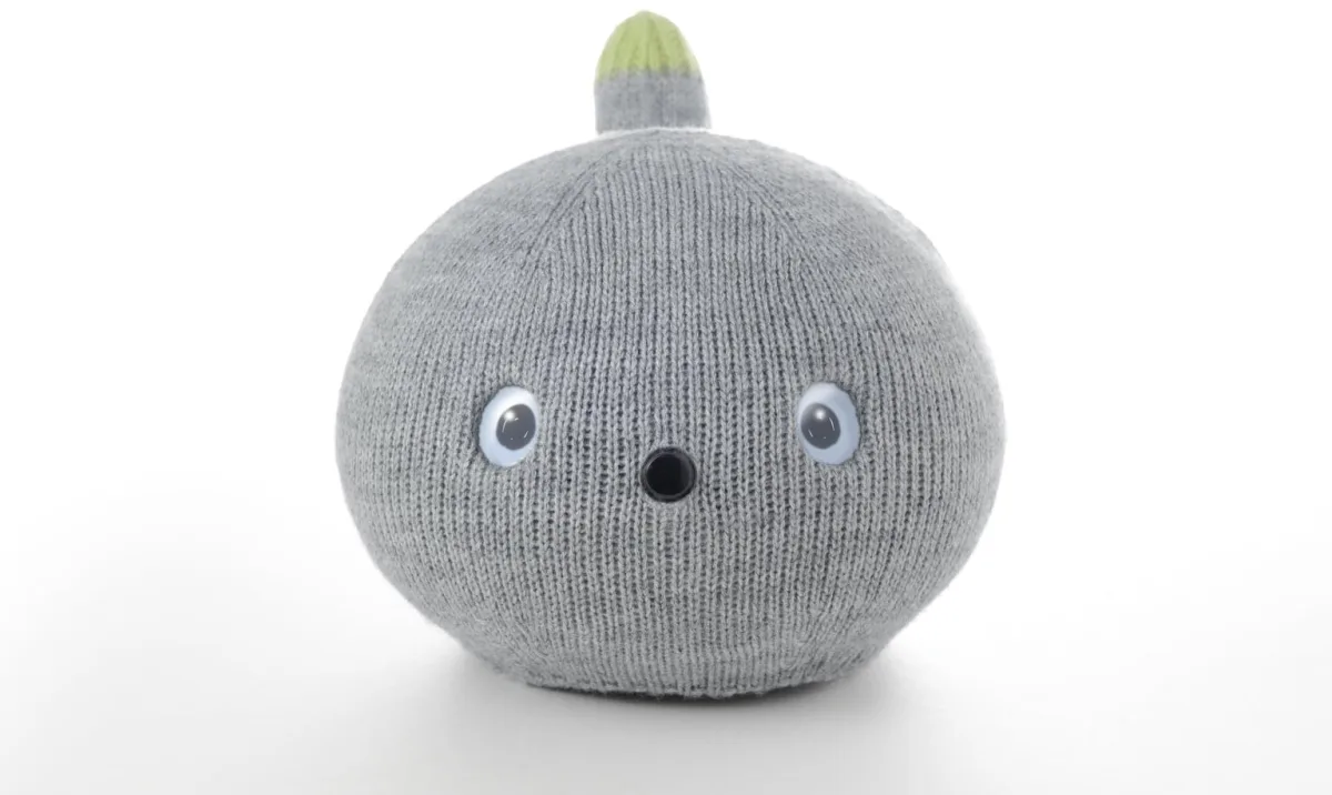 Studio portrait of NICOBO, a small grey companion robot with simple eyes and a sprout-like tail.